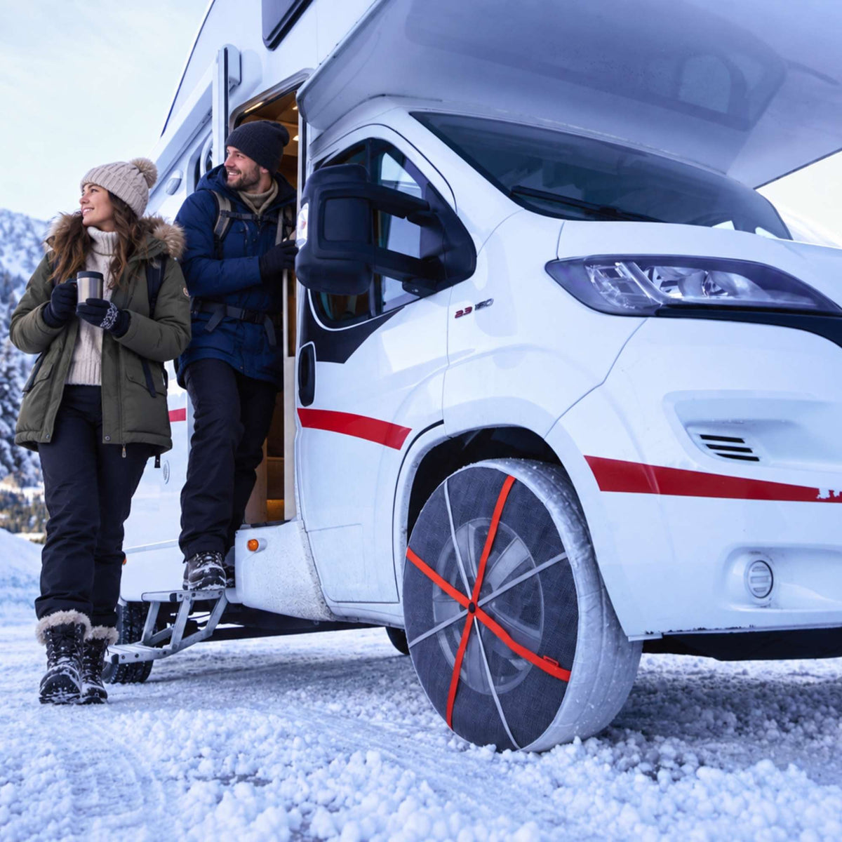 En hvit bobil parkert i snøen med to personer ved den åpne sidedøren. Fordekket er utstyrt med snøsokker med oransje kryssende stropper.